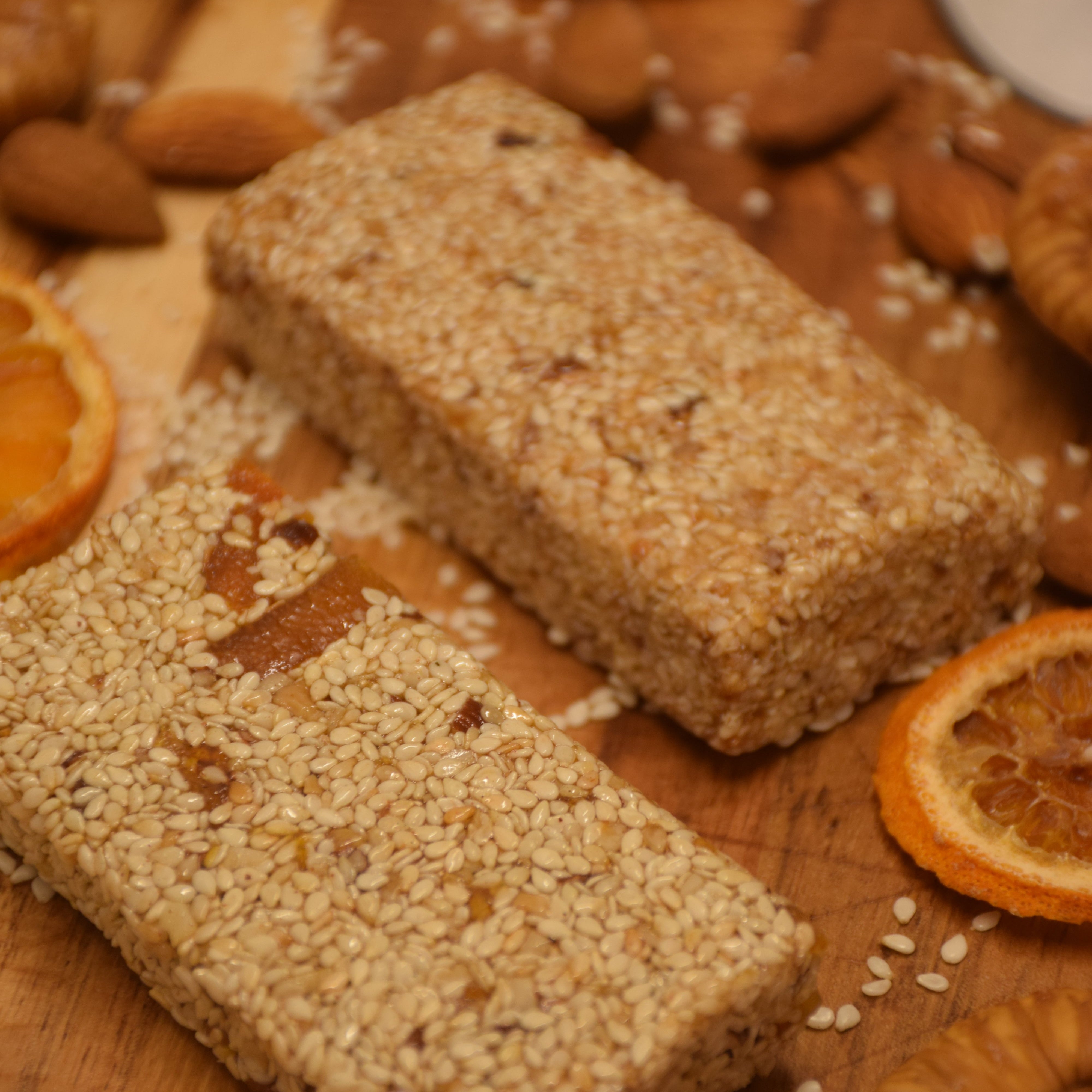Two sesame seed pasteli bars on a wooden board with dried oranges and seeds.
